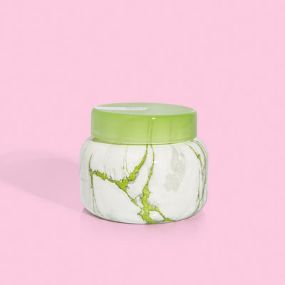 The Honeydew Crush Modern Marble Signature Jar Candle by Capri Blue features a round design with a green lid and marbled white and green pattern, beautifully set against a pink background.