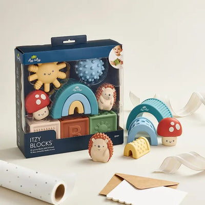 Itzy Ritzys Itzy Blocks features a set of colorful silicon blocks shaped like mushrooms, a rainbow, a sun, and hedgehogs. Loose blocks and ribbon on a beige surface offer sensory textures ideal for creative storytelling.