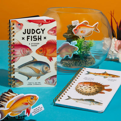 The Judgy Fish Sticker Book by Chronicle Books, a spiral-bound adult sticker book, sits beside a fishbowl of vintage stickers and an open notebook with fish-themed illustrations on turquoise, all set against an orange backdrop.