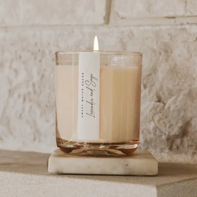 A Sweet Water Decor Lavender and Sage 12 oz Soy Candle in a transparent glass rests on a marble coaster, filling the air with calming French Lavender scent. The stone wall backdrop enhances the serene scene, and the label promises long burn time.