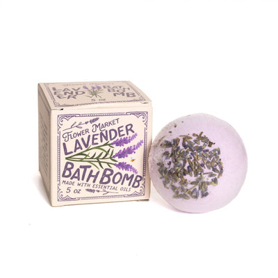A 123 Farm Lavender Bath Bomb - 5 oz, topped with dried lavender, sits next to its box adorned with lavender artwork—ideal for melting away stress and soothing your senses.