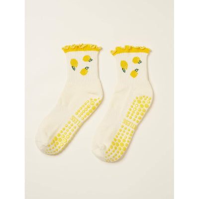 The Lemon Ruffle Grip Sock by Effortless Club is a white organic cotton sock with yellow ruffled cuffs, lemon patterns, and yellow non-slip grips. These Pilates socks feature a cushioned footbed for extra comfort.