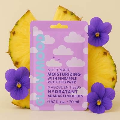 A purple Glow Mood Moisturizing Sheet Mask with Pineapple & Violet Flower stands upright among violet flowers and pineapple slices. The vegan, cruelty-free night mask features cloud designs and is set against a light yellow background.