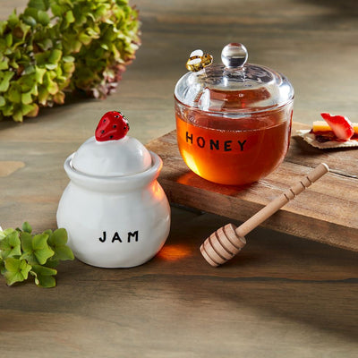 On a wooden surface, the Mud Pie Honey & Jam Set features a stoneware jar with a hand-painted strawberry lid and a glass honey jar with an included spoon. Green leaves in the background enhance the rustic charm.