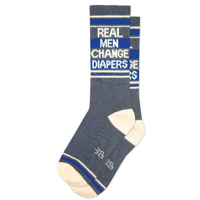 The Real Men Change Diapers Gym Crew Socks by Gumball Poodle feature blue fabric, cream toes and heels, white and blue stripes at the top, and bold text. Made in the USA for comfort and style.