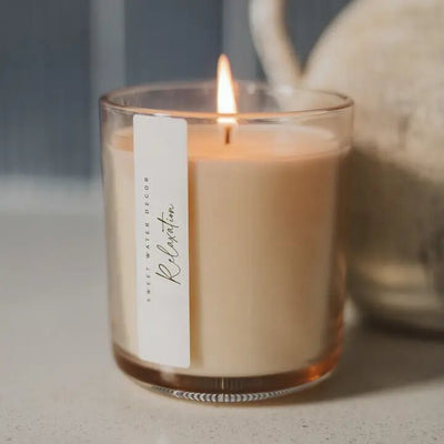 A clear glass jar holds the Relaxation 12 oz Soy Candle by Sweet Water Decor, emitting hints of eucalyptus on a light surface. A blurred object is in the background as the candle casts a warm glow.