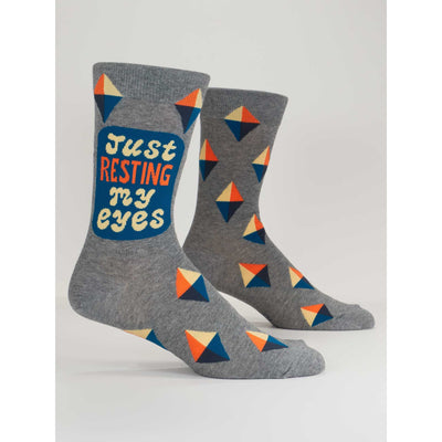 Blue Q Resting My Eyes Mens Socks feature bold geometric patterns in blue, orange, yellow, and white. One sock displays Just resting my eyes in colorful lettering. Fun novelty socks for men and a great addition to your collection!.