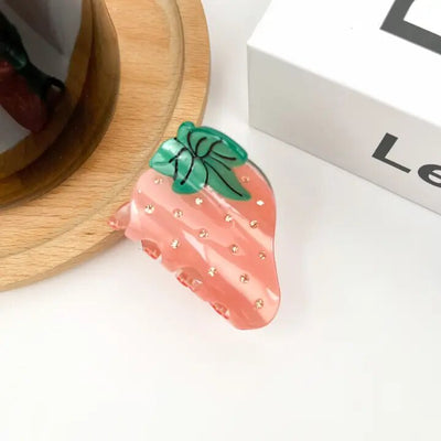 The Diva Soaps Sweet Strawberry Hair Claw Clip, a pink acrylic clip with green leaves and gold dots, rests on a white surface beside a wooden coaster and a white box.