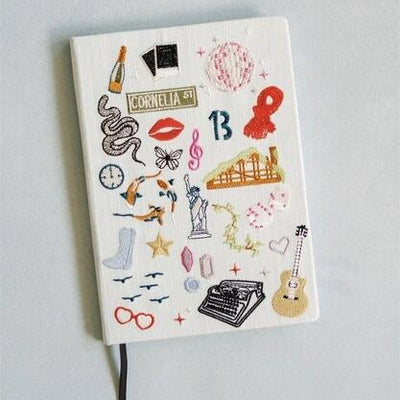 The Gracefully Made Embroidered Taylor Swift Inspired Hardcover Notebook features a linen cover with embroidered icons such as a champagne bottle, snake, lips, musical notes, heart-shaped glasses, and CORNELIA ST, ideal for stylish Swifties to capture their thoughts.