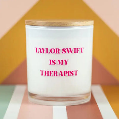 The Taylor Swift Is My Therapist soy candle by Unplug Soy Candles is white with a wooden lid, featuring bold pink text against a geometric backdrop in orange, peach, and green. Perfect for any Swiftie!.