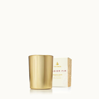 A Thymes Frasier Gilded Gold Votive Candle stands beside its white box, adding a touch of forest fragrance—an ideal hostess gift—against a plain, light background.