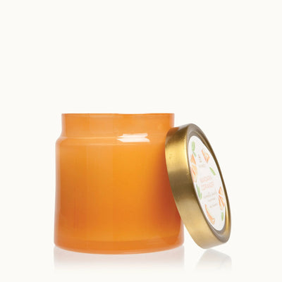 An orange jar with a gold lid, slightly open, reveals Mandarin Coriander Statement Poured Candles contents by Thymes. The lid showcases an illustrated label with orange slices against a plain white background.