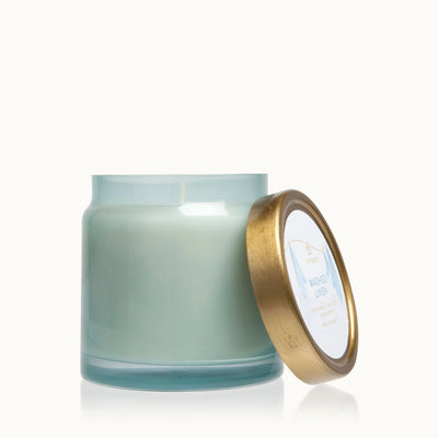 A Washed Linen Statement Poured Candle by Thymes in light blue, infused with a calming scent, rests in a transparent glass jar. Its gold lid is slightly open, revealing the label. The candle is set against a plain white background.