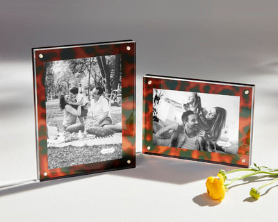 Two black and white photos of cheerful people are showcased in Mud Pies Tortoise Block Frames, one vertical and one horizontal. They rest on a sunlit surface beside two small yellow flowers. The acrylic construction lends a modern touch to the vintage aesthetic.