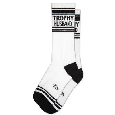 The Gumball Poodle Trophy Husband Unisex Socks are white mid-calf socks with black toe and heel accents, black stripes near the top, and TROPHY HUSBAND boldly written around the upper section.