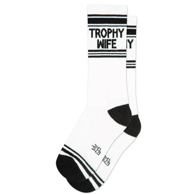 Gumball Poodle’s Trophy Wife Gym Crew Socks are white unisex mid-calf socks with black toes, heels, and stripes, featuring “TROPHY WIFE” in bold black letters near the top. The wide ribbed socks are shown flat on a white background.