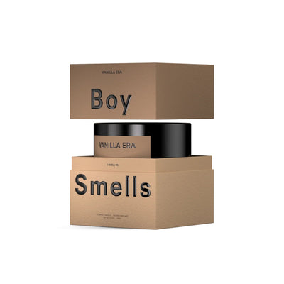 A brown rectangular Boy Smells box sits beside a black cylindrical Vanilla Era Hero Candle, labeled with scents of warm vanilla and golden amber, shown against a white background. Brand: Boy Smells.