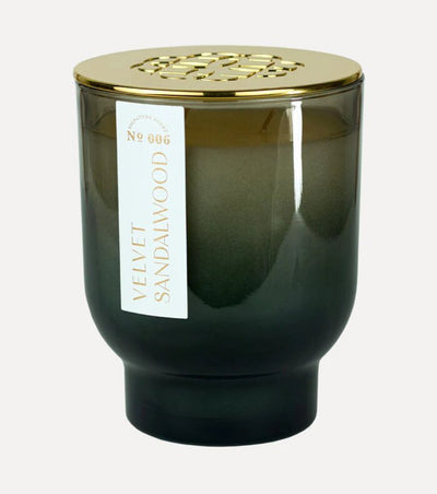 The 21 oz Maeve Glass Candle by Elum, in a green gradient design labeled Velvet Sandalwood, is adorned with a decorative gold lid featuring intricate cut-out patterns. The white and gold text on its vertical label enhances its elegance while exuding serenity and relaxation.