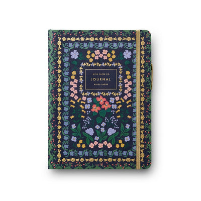 The Wildwood Journal with Pen by Rifle Paper Co. is a hardcover adorned with vibrant floral patterns. It features a central dark square labeled Journal, secured by a yellow elastic band, and contains ruled pages perfect for pairing with your favorite gold pen.