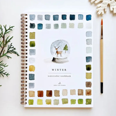 The Emily Lex Studio Winter Watercolor Workbook, spiral-bound for beginners, rests on a white surface. Its cover displays wintery color swatches, a snow globe with a reindeer, a paintbrush, and a pine branch.