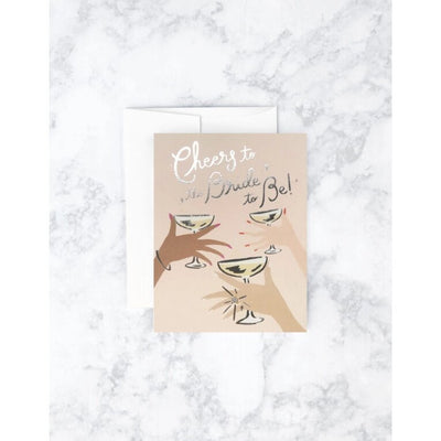 On a marble surface, the Idlewild Co. Cheers to the Bride Card features illustrated hands holding champagne glasses with the text Cheers to the Bride to Be! Ideal for bachelorette celebrations, it comes with a partially visible envelope.