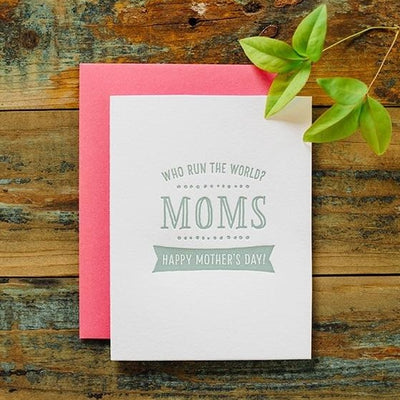 The Harken Press Moms Run the World Greeting Card sits on a wooden surface with a pink envelope. It features Who run the world? MOMS, and Happy Mothers Day! in a banner underneath, while a green leafy branch decorates the corner for a natural touch.