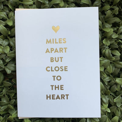 The Miles Apart Greeting Card by Sugar Paper features a card with the message Miles apart but close to the heart in elegant matte gold foil, topped with a delicate gold heart, all set on a bed of green leaves.